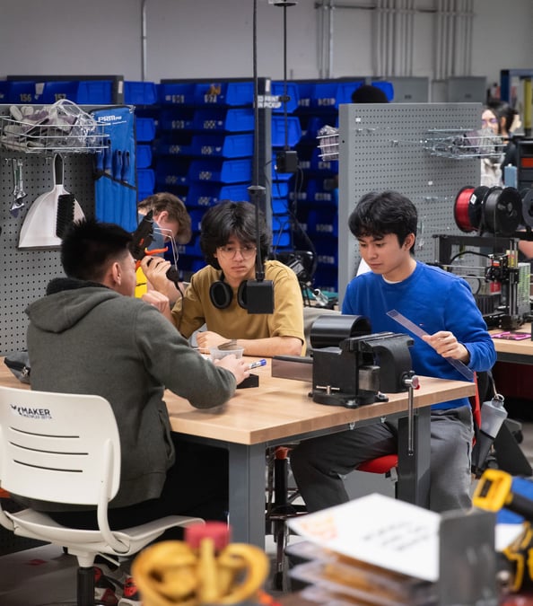 Studenci przy stole warsztatowym w makerspace Schulich, za nimi pracujące drukarki Prusa