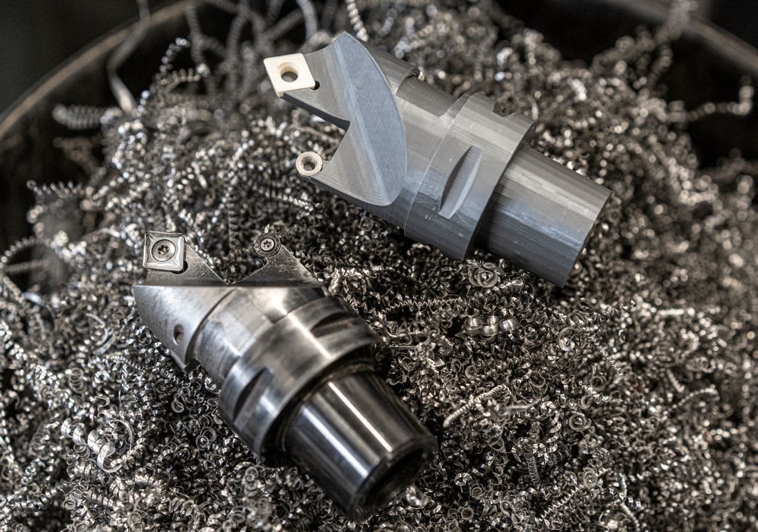 A grey 3D printed model of a combined cutting tool with a white insert fitted in its seat, lying beside the finished steel production tool on a bed of curled steel swarf