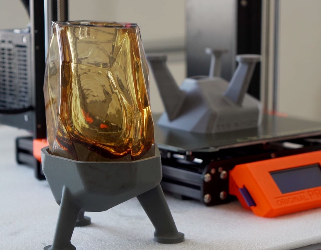 An amber blown glass lamp shade seated in a grey 3D printed tripod base, with an Original Prusa printer behind it