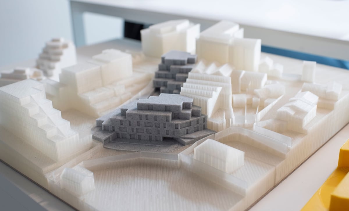 A 3D-printed urban massing model on a light board: translucent white printed building volumes with two buildings picked out in grey