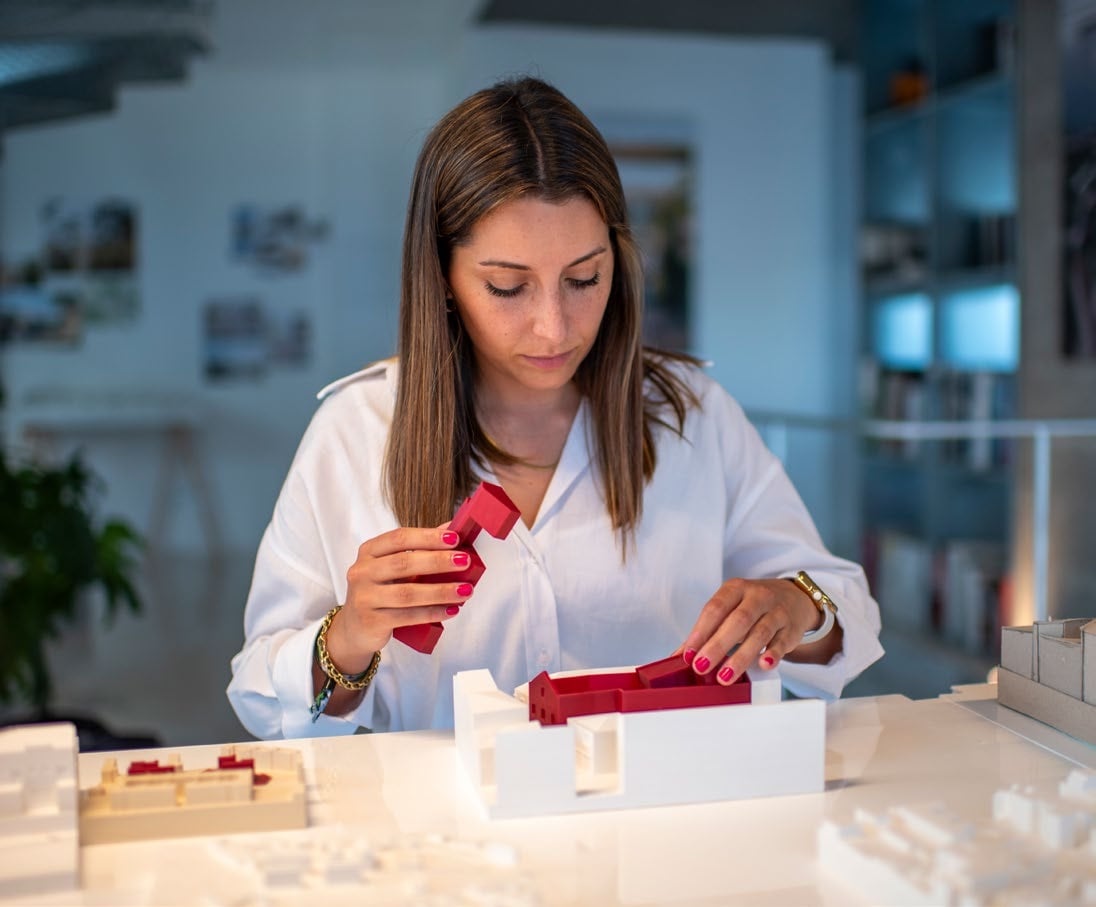 An architect at a studio table lifting a red 3D-printed building volume while seating a second red piece into a white printed base model