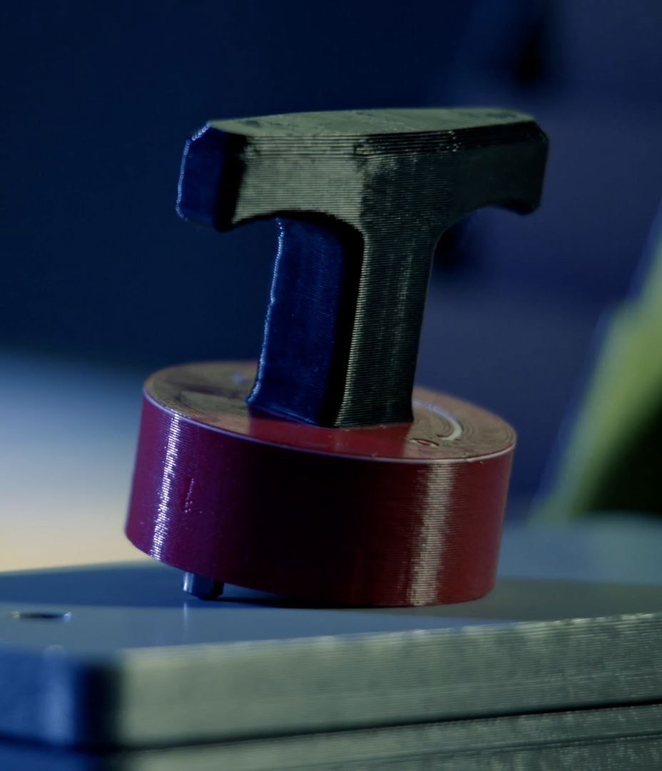 A small black 3D printed T handle on a dark red printed cylindrical base, standing on a machined metal plate, layer lines visible on both parts