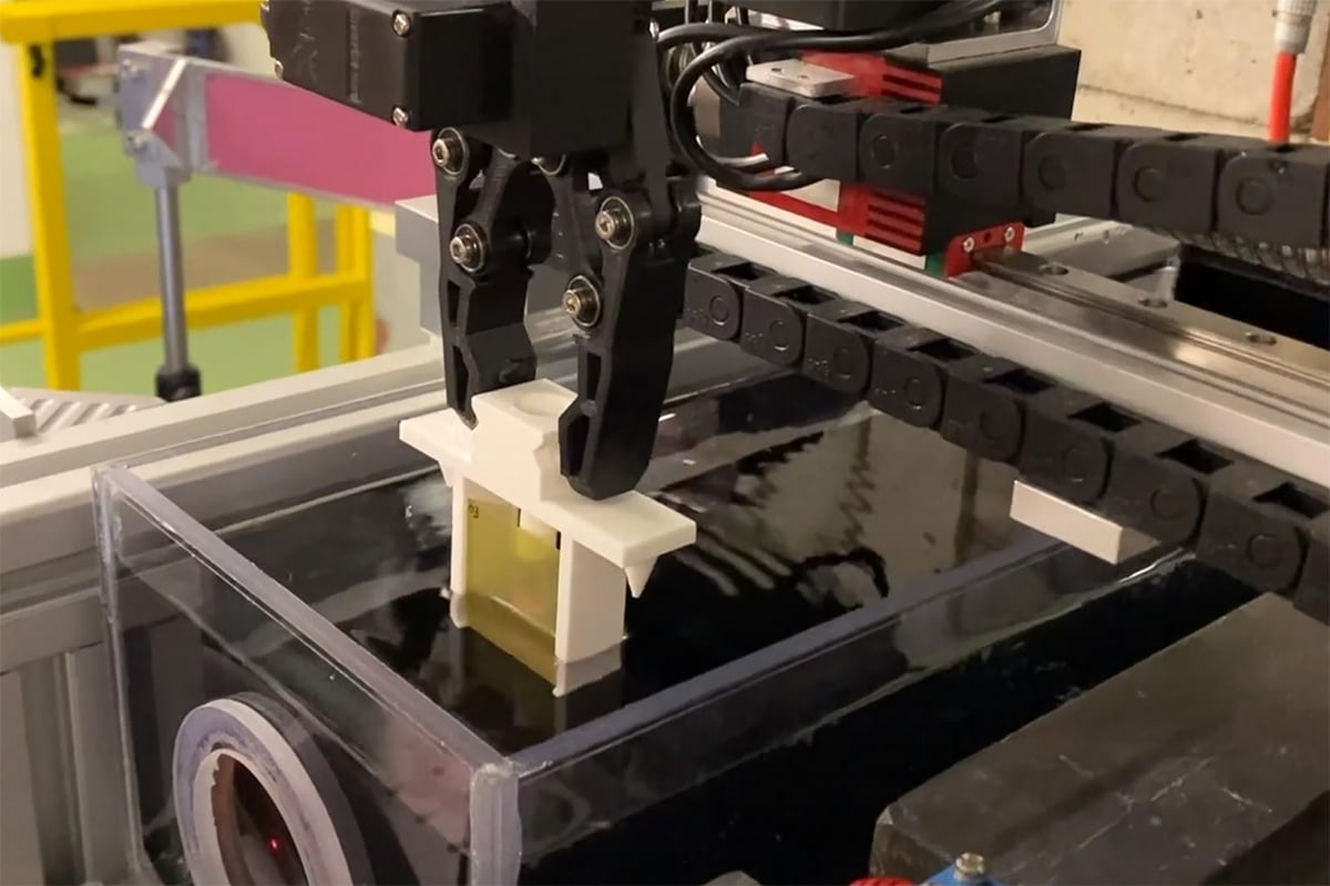 A black printed two-finger gripper on a linear rail lowering a white printed sample holder into a clear tank