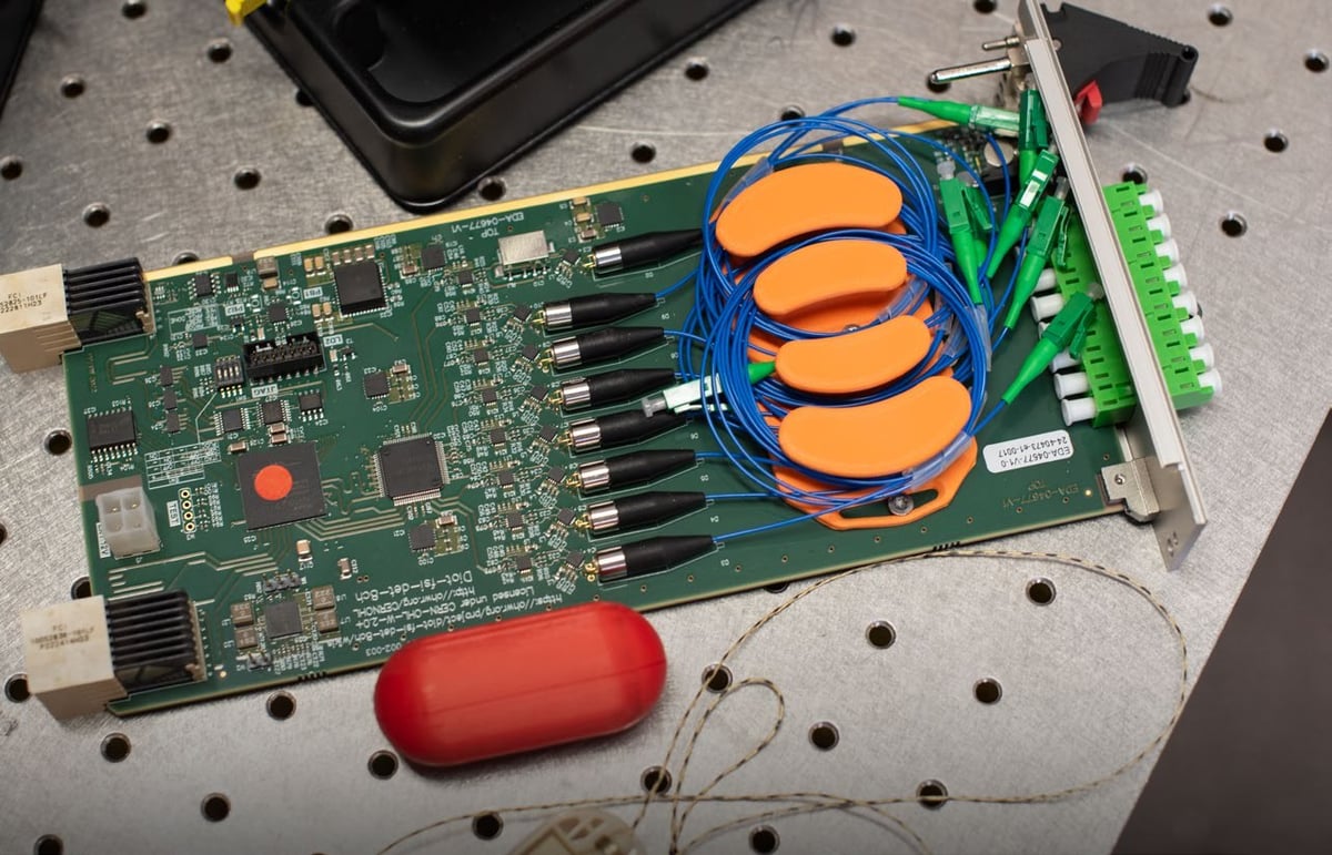 A green circuit board with four orange printed spools holding coiled blue fibre-optic cable, and a red printed capsule beside it
