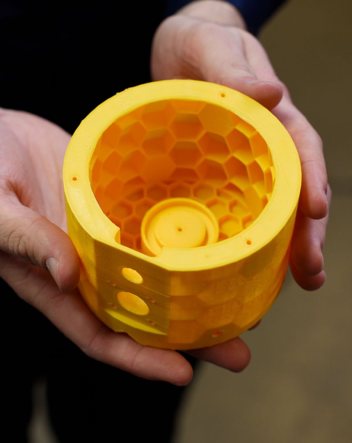 Two hands holding a yellow printed cylindrical part with a honeycomb interior