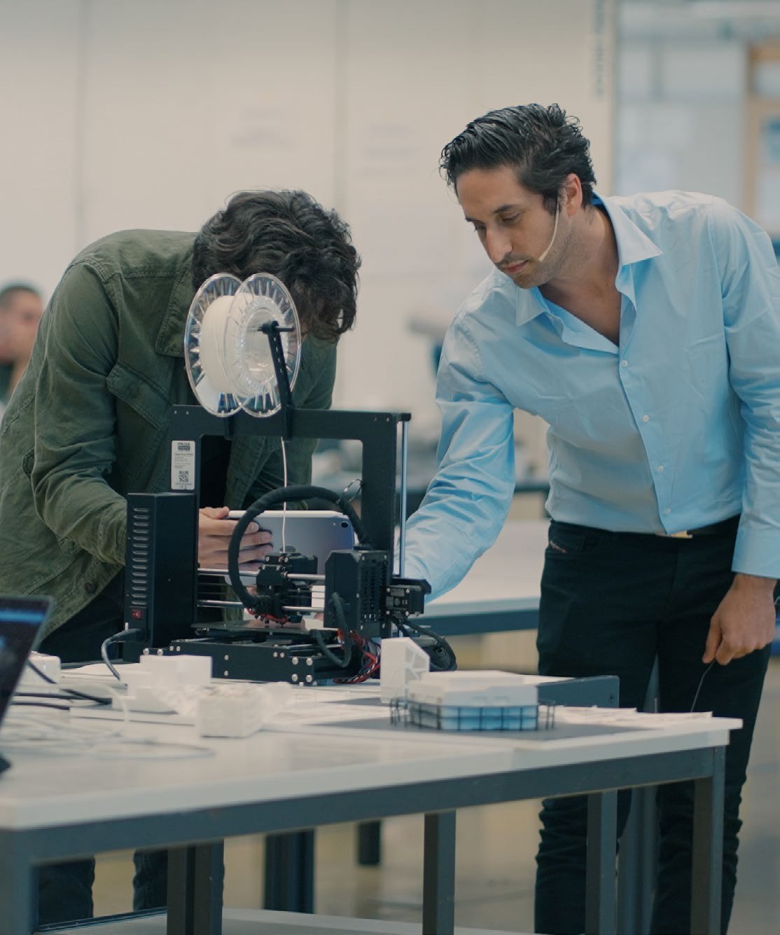 Jonathan Benhamu leaning over an Original Prusa printer beside a student, printed model pieces on the table