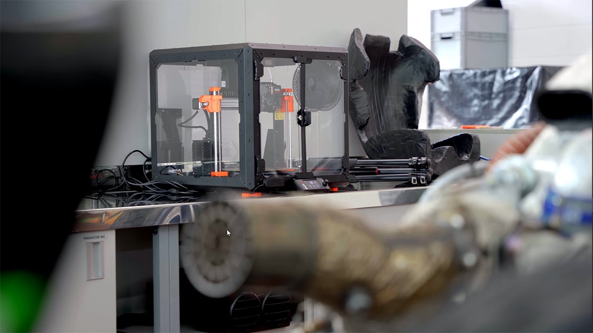 An Original Prusa MK4 on a flight case printing a black airflow manifold, beside the AIX single seater in green and bronze livery, car number 25
