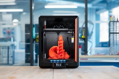 A Prusa CORE One+ printing an orange model on a bench in a bright workshop