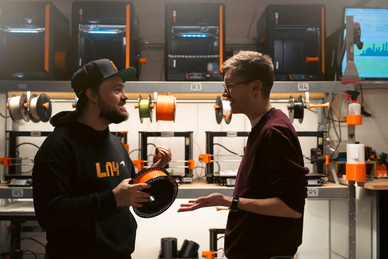 A man showing a Prusament spool to another colleague in a 3D printer farm setting.